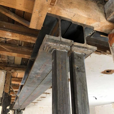 Zwei metallene I-Balken sind an einem Holzrahmen befestigt, wobei ein Teil des Balkens in einer Vertikalen und der andere in einer Horizontalen Position zu sehen ist.